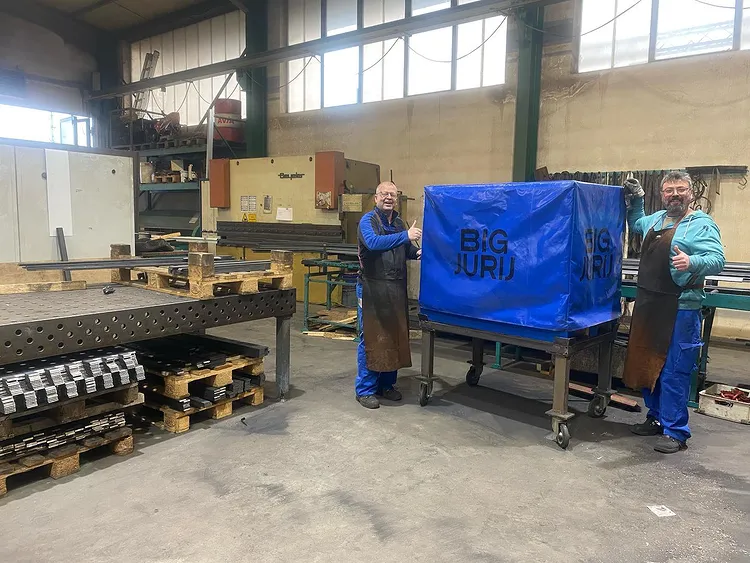 Zwei Arbeiter in einer Werkhalle stehen neben einem blauen Tisch mit der Aufschrift "BIG JURIJ" und zeigen Daumen hoch.